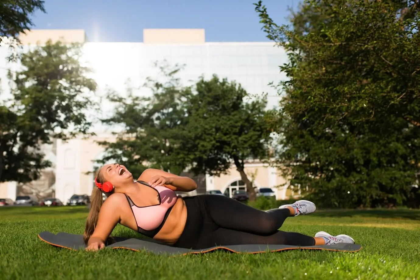 Jonge lachende plus-size vrouw in een sportieve top en legging met rode koptelefoon, liggend op een yogamat terwijl ze vrolijk tijd doorbrengt in een stadspark.