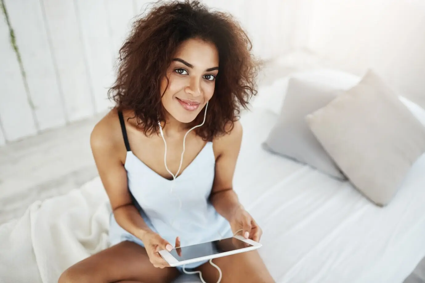 Een mooie Afrikaanse vrouw in slaapkleding zit op bed, glimlacht en luistert met een koptelefoon naar muziek.