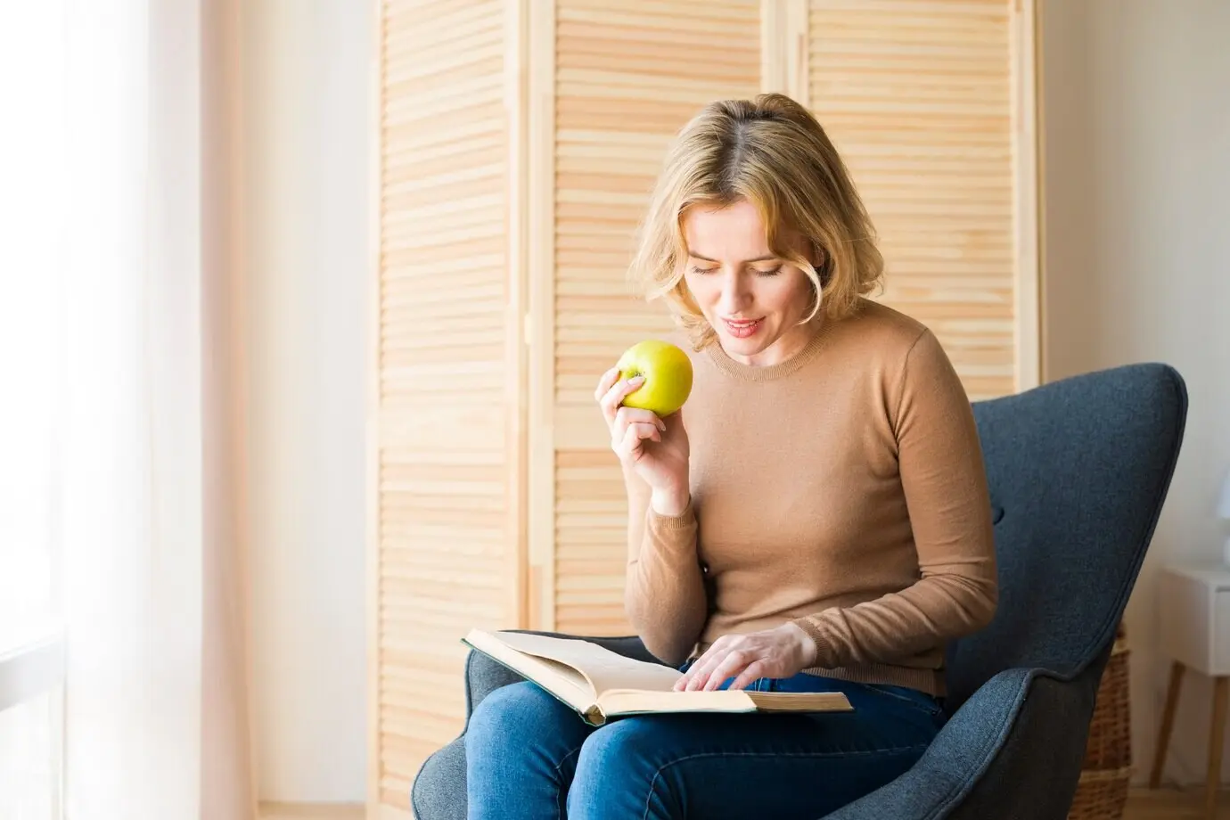 Een blonde vrouw leest een boek terwijl ze een appel eet.