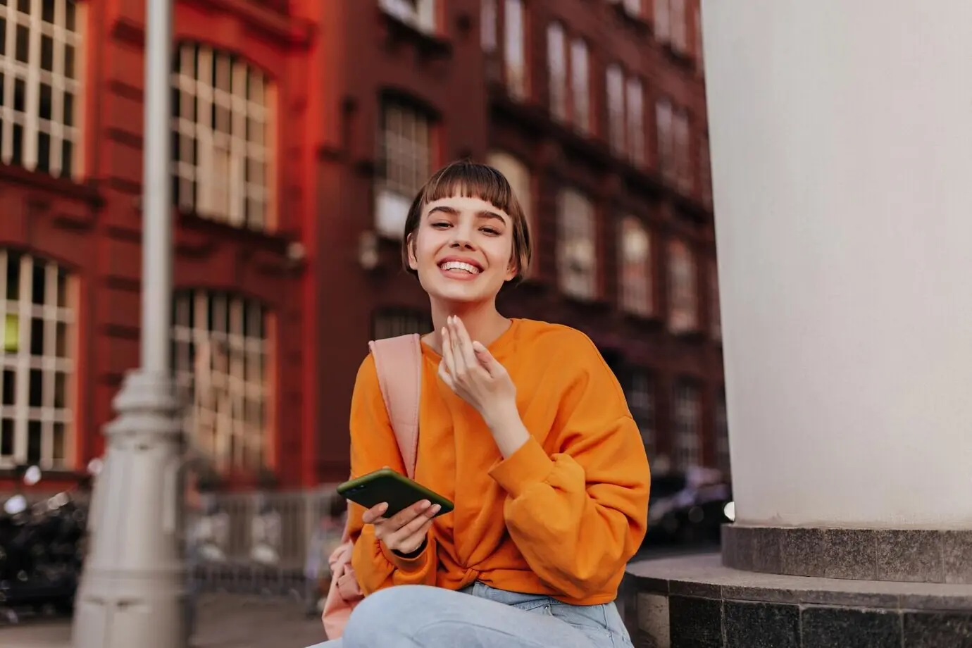 Jonge vrouw met kort haar in een oranje sweatshirt glimlacht oprecht buiten. Blije, coole meid houdt een telefoon vast en zit buiten.
